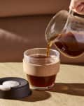Pouring coffee from a glass carafe into a cork-wrapped glass cup on a wooden table.