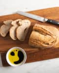 Sliced bread on a wooden cutting board with a serrated knife and a bowl of olive oil and balsamic vinegar.