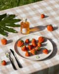Strawberries on a plate with a bottle of honey on a checkered tablecloth, surrounded by leaves and flowers.