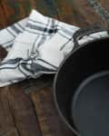 Cast iron Dutch oven with lid and brass handle on rustic wooden table, next to a black and white checkered cloth.