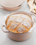 Freshly baked bread in a pink Dutch oven on a marble countertop, with butter and herbs on a tray in the background.