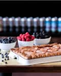 Baked pie in a white dish on a wooden table with bowls of blueberries, raspberries, and blackberries.