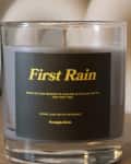 Glass candle labeled "First Rain" with scent description "Rain on the Pavement" on a wooden surface.