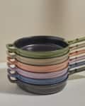 Stack of colorful frying pans with handles in green, beige, pink, blue, and black on a light surface.