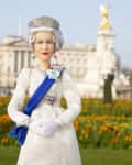 A doll in a white gown with a blue sash stands in front of Buckingham Palace, surrounded by orange flowers.