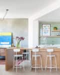 Modern kitchen with light wood island, white stools, green tile backsplash, and open shelves with decor and plants.