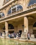 Visitors exploring the Roman Baths, featuring a green pool, classical architecture, and stone columns.