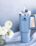 Blue Stanley mug with handle and straw on striped cloth, next to white flowers and kitchen items.