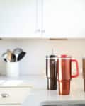 Three Stanley tumblers in black, red, and gold on a white kitchen counter with utensils in the background.
