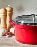 Red Dutch oven with glass lid on wooden table, surrounded by garlic, pepper mills, and olive oil can.
