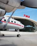 Vintage TWA airplane parked outside the TWA terminal with curved glass facade and red TWA sign.