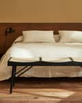 Minimalist bedroom with a white bed, wooden headboard, black wall lamp, and woven bench on wooden floor.