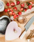 Heart-shaped chocolate cake dusted with powdered sugar, surrounded by fresh strawberries, a knife, and baking tools on a wooden surface.