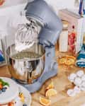 Blue stand mixer with whipped cream, surrounded by a cake with fruit, orange slices, eggshells, and cookbooks.