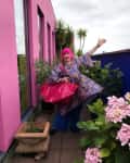 Person with pink hair in colorful attire poses on a balcony with pink walls, surrounded by potted plants and flowers.