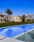 Modern luxury home with large glass windows, palm trees, and a blue swimming pool reflecting the evening sky.