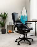 Home office with black swivel chair, wooden desk, laptop, potted plant, surfboard, and large window with blinds.