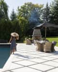 Woman in a sun hat walking by a pool, with patio furniture and a fireplace under a black umbrella in a garden setting.
