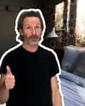 Man giving thumbs up in a bedroom with floral wallpaper, gray bedding, and a wall lamp.