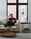 Man sitting on a cream sofa with pillows, next to a round coffee table and a floor lamp, in a modern living room.