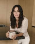 Woman with long dark hair holding a mug, leaning on a kitchen counter.