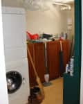 Cluttered laundry room with stacked washer-dryer, wooden cabinets, broom, and cleaning supplies.