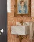 A powder room with orange wallpaper that has white squiggly lines on it, and a marble sink below a framed portrait. The portrait has a gold frame, and the sink has gold hardware. There's a beige towel with a gold towel ring beside it.