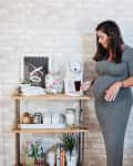 Pregnant woman in gray dress using a coffee maker on a wooden shelf with mugs, jars, and decor against a brick wall.