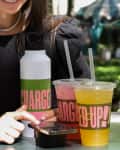 Woman in black dress using smartphone at outdoor table with two colorful drinks and a water bottle labeled "Charged Up!"