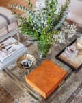 Glass coffee table with a floral arrangement, books, a wooden box, and a tray with a candle and matches on a patterned rug.