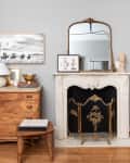 Wooden dresser with lamp and decor beside ornate fireplace with gold screen and mirror.