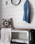 Round mirror and hooks on shiplap wall, denim shirt hanging, bench with patterned pillow and white throw.