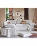 White sectional sofa with abstract pillows, side table, and a large plant in a neutral-toned living room.