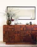 Wooden sideboard with geometric design, large mirror, vase with foliage, and a cane rocking chair with a white fur cushion.