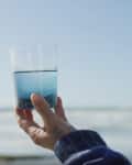 Hand holding a glass of water against a clear blue sky and ocean backdrop.
