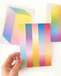 Hand holding a colorful gradient postcard with two abstract gradient art prints in the background.