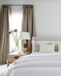 Beige bedroom with a bed, white bedding, two lamps, and matching curtains, flanked by two windows.