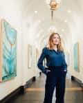 Woman in a blue blouse standing in an art gallery hallway with abstract paintings on the walls.