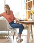 Woman in a coral shirt sitting at a wooden dining table with a vase of yellow flowers, reaching towards a shelf.