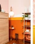 Room with orange and yellow walls, floor lamp, wooden shelves, boots, and framed artwork on floral wallpaper.