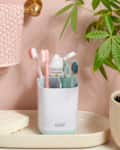 bathroom sink scene with a soap dispenser, lit candle, bar soap, and a white toothbrush organizer with toothbrushes and toothpaste in them against a peach wall with a plant on the right