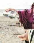 Person pouring water from a bottle into a glass on a sandy beach, wearing a pink and black patterned garment.