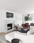 Living room with a marble fireplace, cream sectional sofa, leather armchair, floor lamp, and large potted plant.