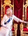 Elderly woman in a white gown and tiara petting a corgi on a red and gold throne with a Union Jack pillow.