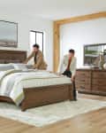 Two men making a bed in a modern bedroom with wooden furniture, striped bedding, and decorative vases on a dresser.