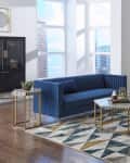 Modern living room with blue velvet sofa, geometric rug, black cabinet, and framed leaf prints on the wall.