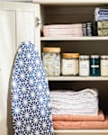 Linen closet with folded towels, labeled jars, cleaning supplies, and an ironing board with a blue geometric cover.