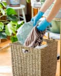 Person wearing blue gloves placing laundry in a wicker basket, surrounded by green plants and a chair with green cushions.