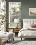 Bedroom with green walls, tufted headboard, wooden nightstands, and large windows overlooking a garden.