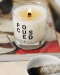 Lit candle labeled "FOCUSED" on a book, next to a bowl with a burnt sage bundle.